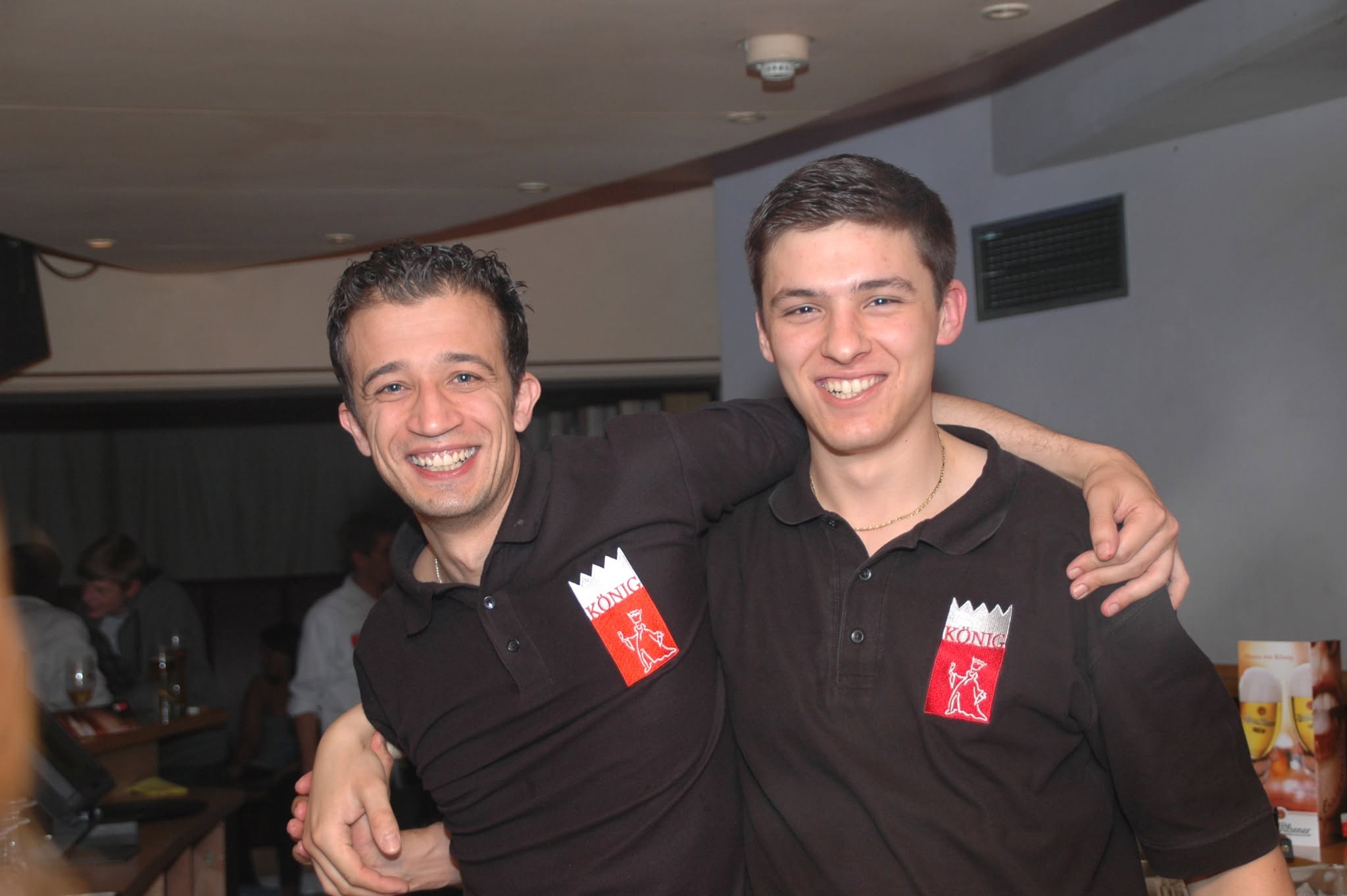 Zwei lächelnde Personen in schwarzen KÖNIG-Polos stehen Arm in Arm.
